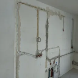 Ściana z bruzdami i ułożonymi przewodami elektrycznymi. Widoczne puszki instalacyjne w kolorze czerwonym. Przewody zabezpieczone uchwytami. Etap prac instalacyjnych.