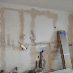 Ściana w trakcie remontu: widoczne instalacje wodne, elektryczne, ślady po kuciu, oznaczenia na ścianie i przygotowanie do dalszych prac wykończeniowych.