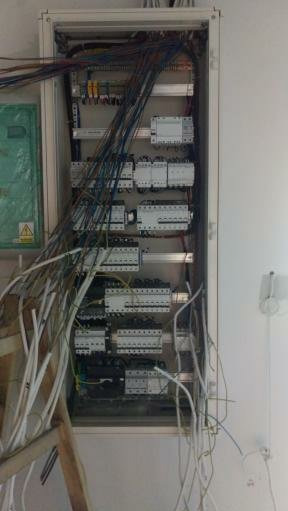 Rozdzielnia elektryczna w trakcie montażu z licznymi przewodami, widoczne zabezpieczenia i elementy sterujące, w tle fragment ściany i drabina.