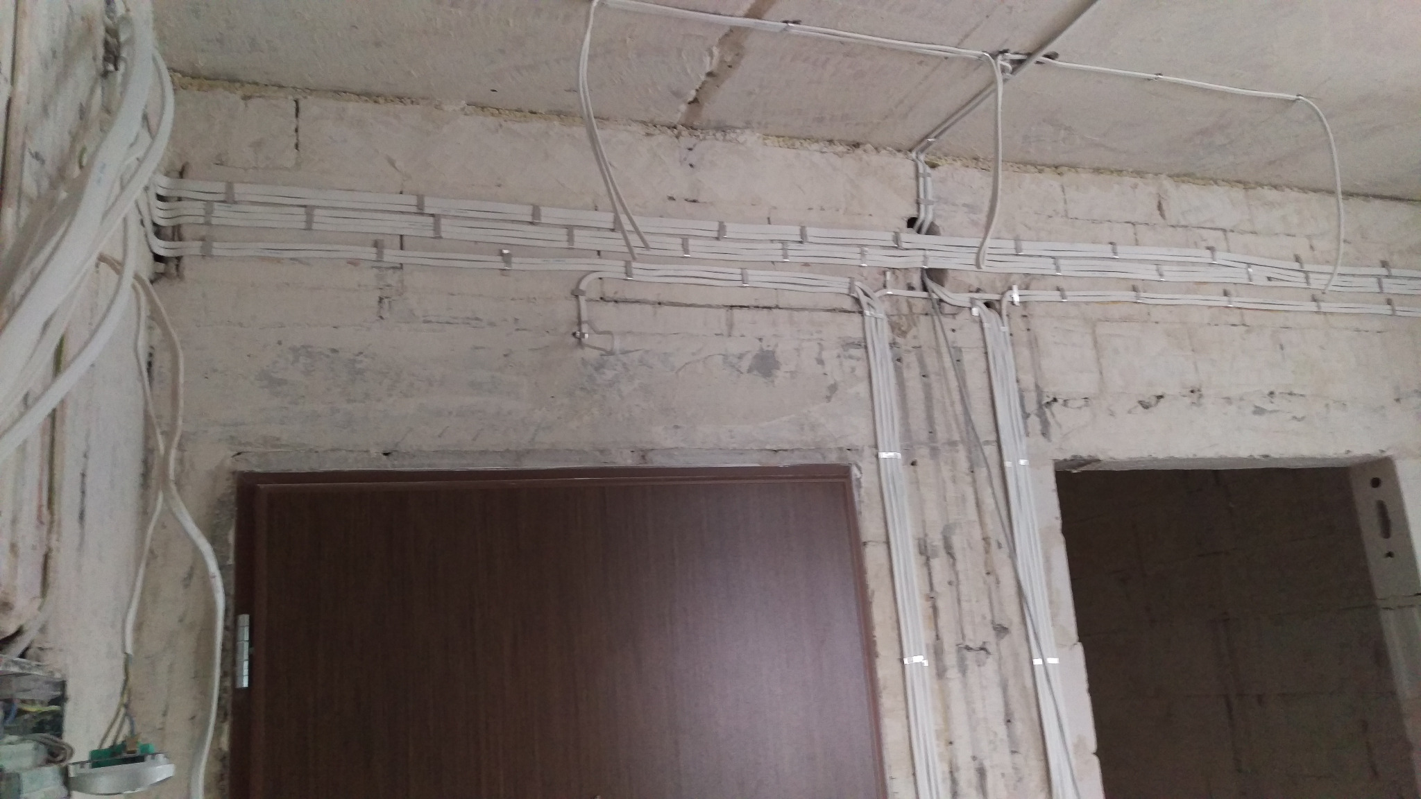 Białe kable elektryczne ułożone na ścianie z betonu komórkowego nad brązowymi drzwiami i otworem drzwiowym. Instalacja zabezpieczona metalowymi uchwytami.