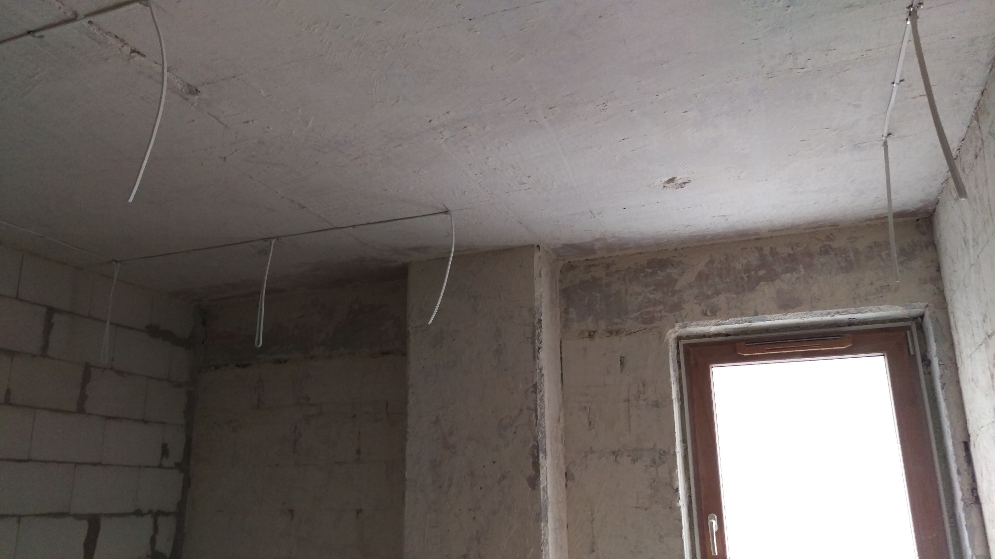 Wnętrze w trakcie prac: surowy strop z kablami elektrycznymi, ściany z betonu komórkowego i okno. Widoczne przygotowania do instalacji oświetlenia.