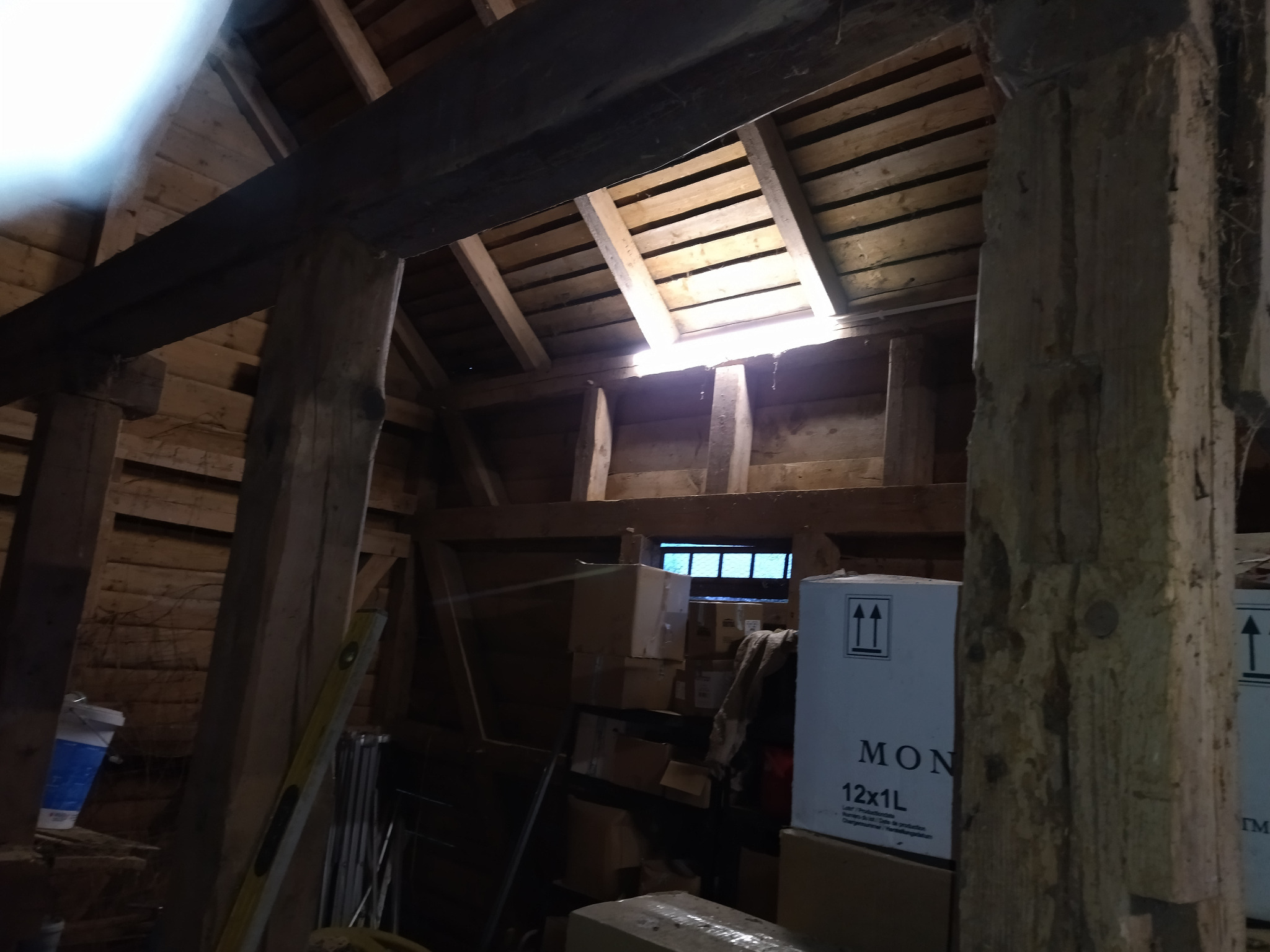 Drewniana konstrukcja dachu z widocznym oknem dachowym i elementami instalacji, w tle kartony i narzędzia. Ujęcie z dołu, ukazujące detale konstrukcyjne.