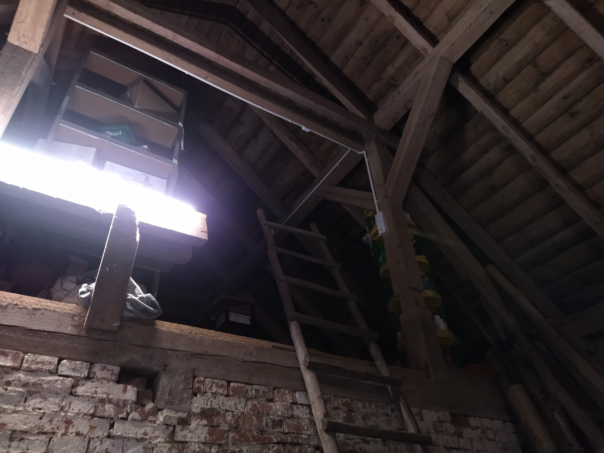 Strych z drewnianą konstrukcją dachu i widoczną białą instalacją elektryczną biegnącą wzdłuż belek. Drewniana drabina oparta o ceglaną ścianę.