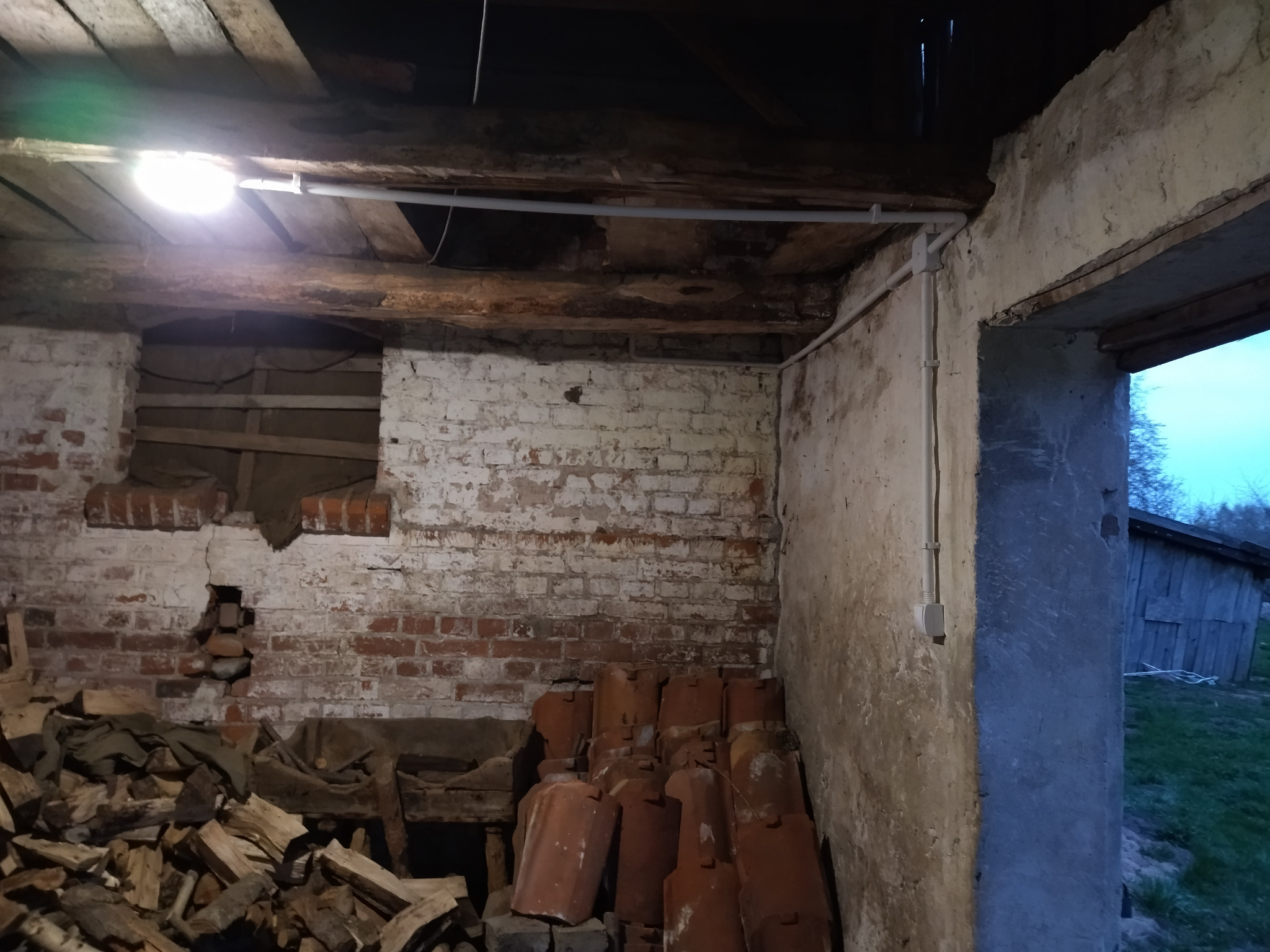 Wnętrze starego budynku z odsłoniętą cegłą, oświetlone lampą. Widoczne przewody elektryczne i elementy instalacji. Część pomieszczenia zagracona drewnem i dachówkami.