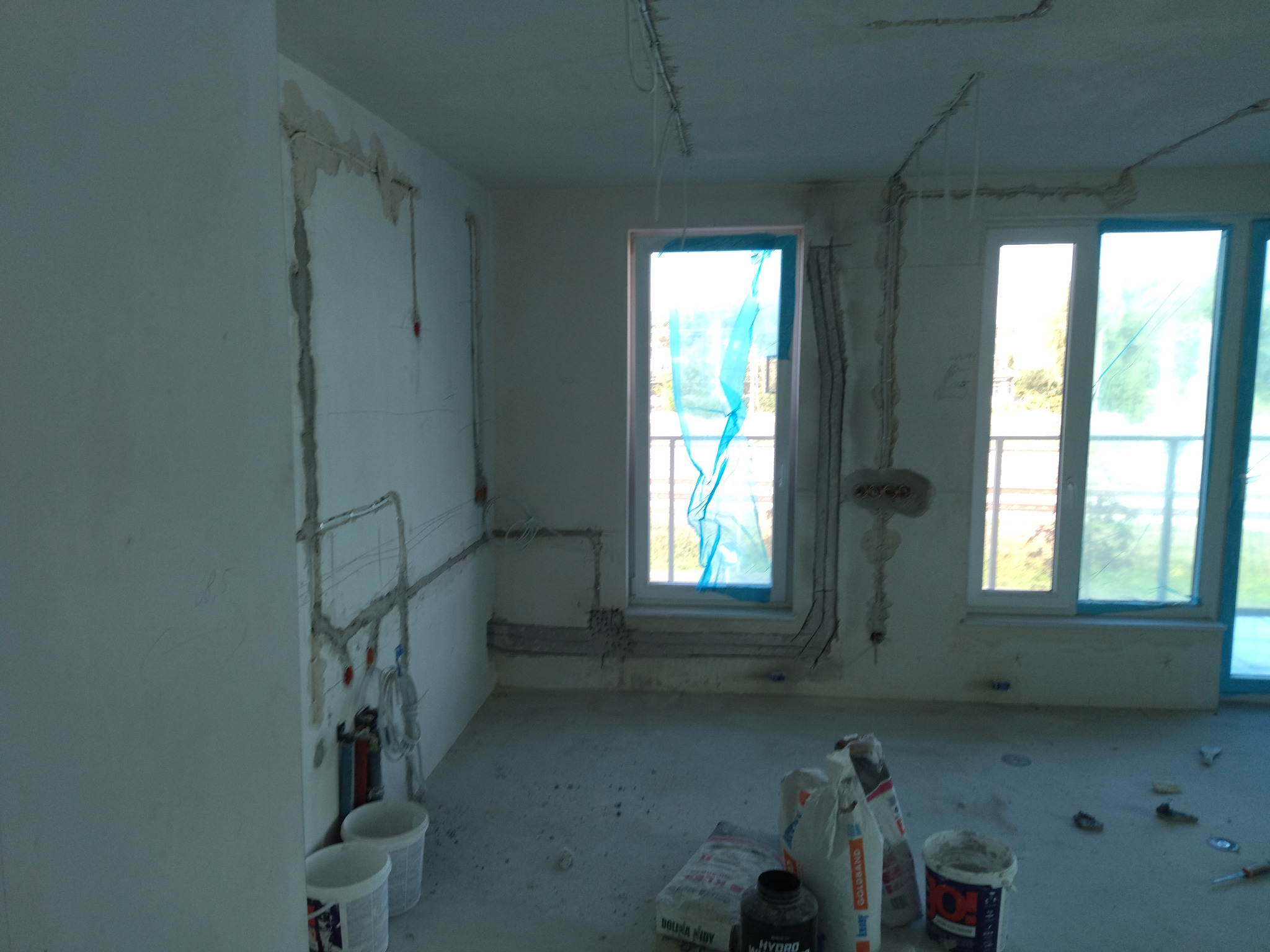 Wnętrze w trakcie remontu: widoczne kable elektryczne w ścianach, worki z zaprawą i puste wiadra. Okna zabezpieczone folią. Etap prac instalacyjnych.