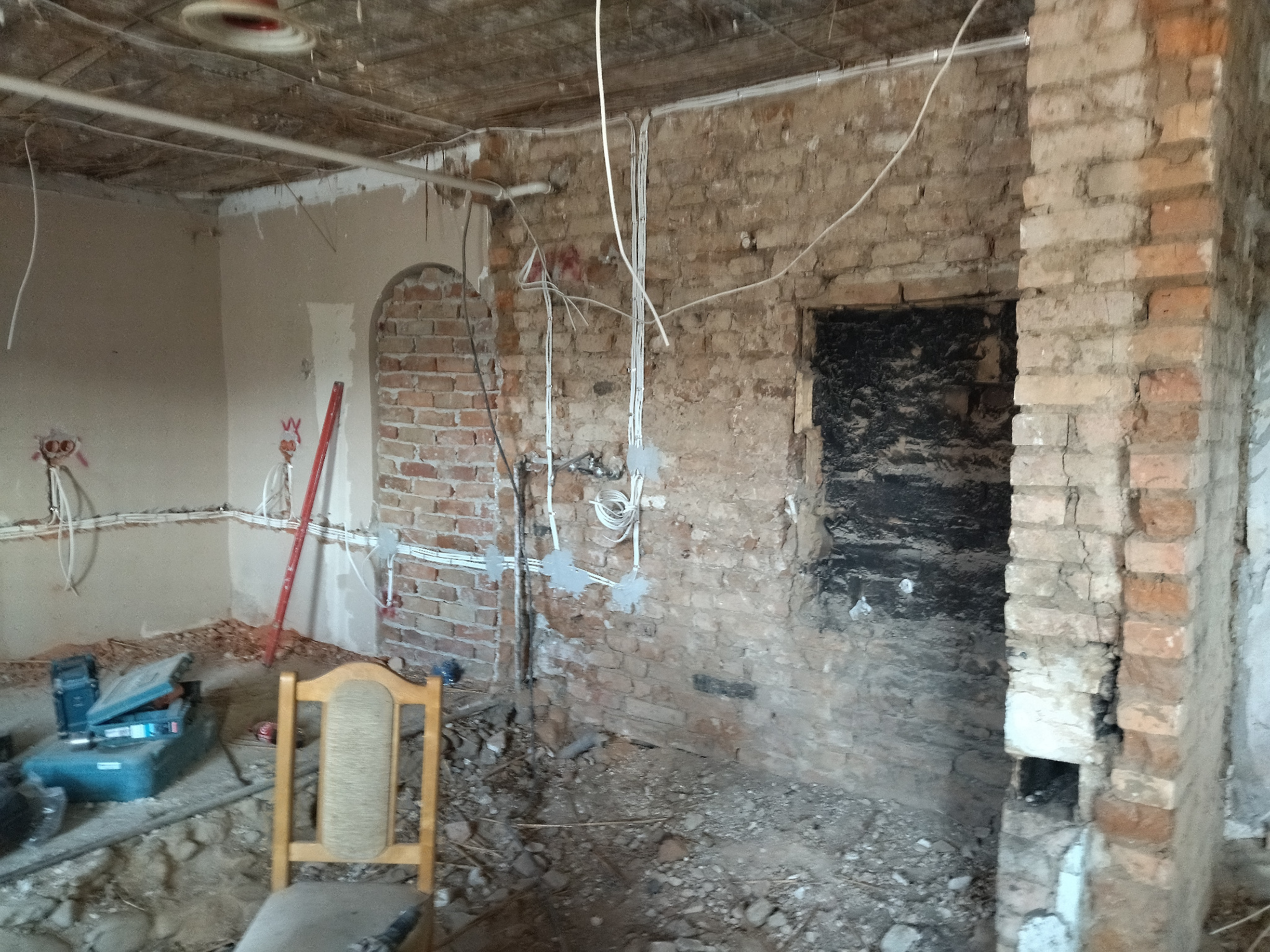 Wnętrze w trakcie remontu: odsłonięta cegła, kable elektryczne w ścianach, narzędzia i poziomica. Widoczne ślady po kuciu i wierceniu. Krzesło w pomieszczeniu.