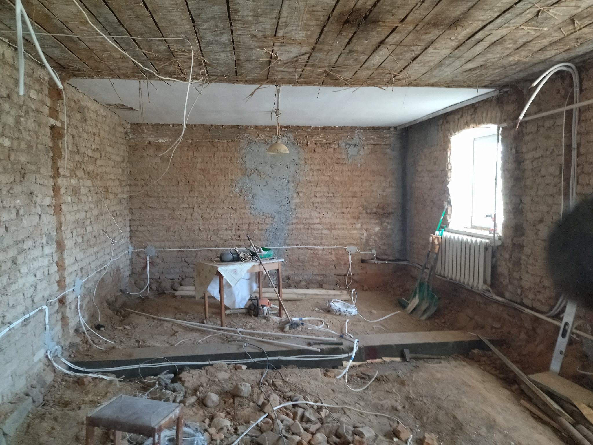 Wnętrze pokoju w trakcie remontu z odsłoniętą cegłą, widocznymi przewodami elektrycznymi w białych rurkach i prowizorycznym oświetleniem.