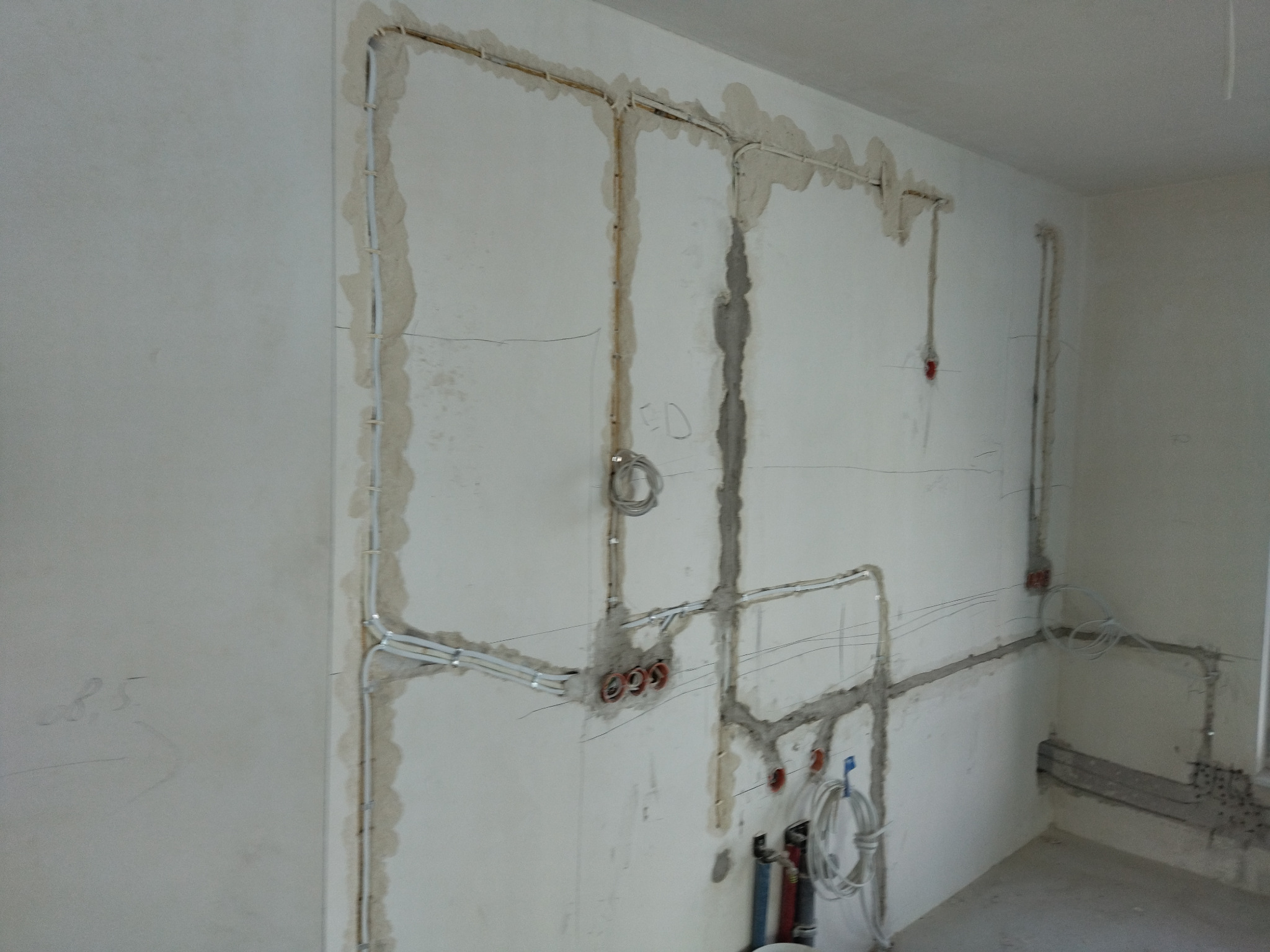 Ściana z bruzdami i ułożonymi przewodami elektrycznymi. Widoczne puszki instalacyjne w kolorze czerwonym. Przewody zabezpieczone uchwytami. Etap prac instalacyjnych.