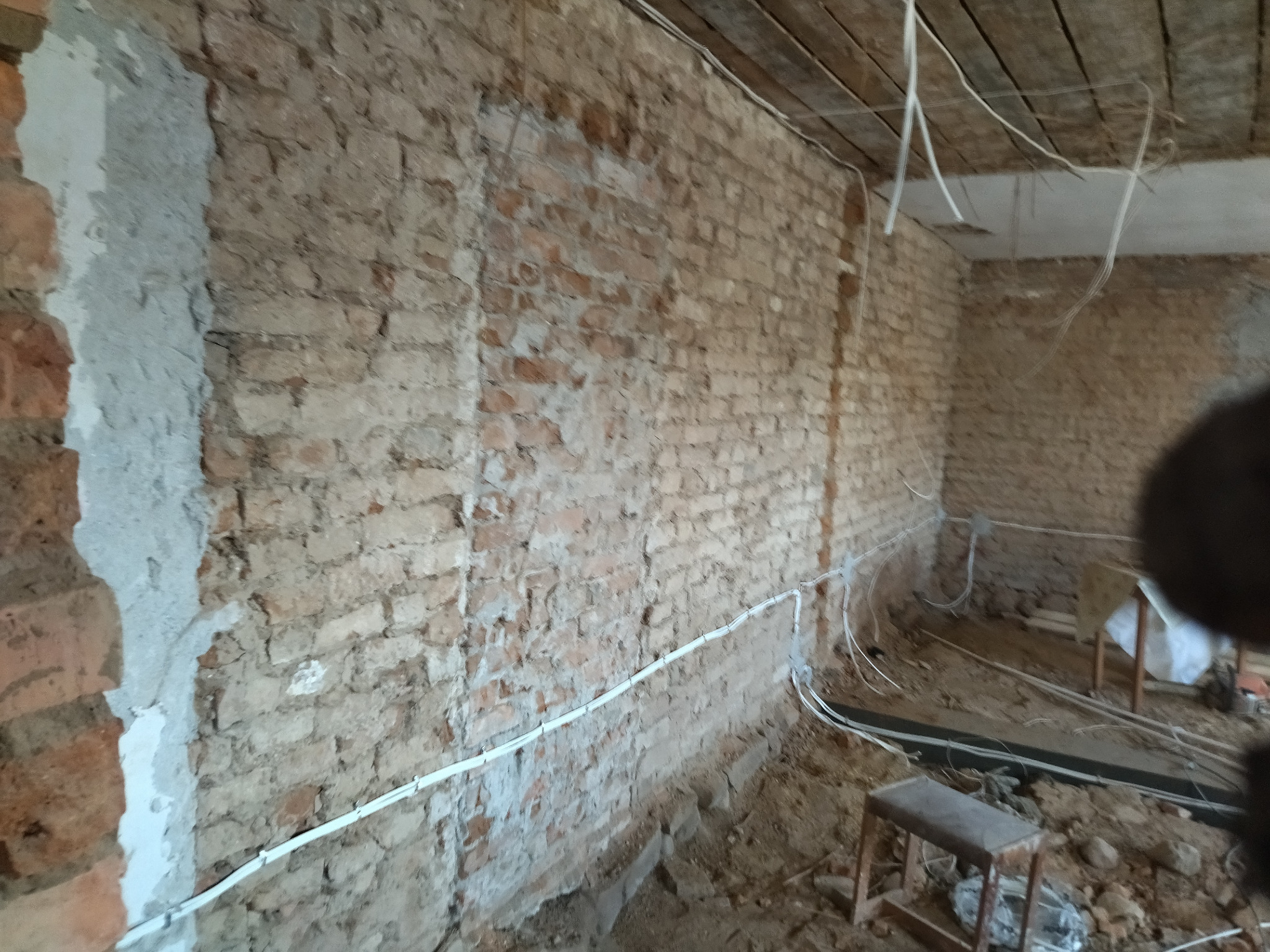 Ceglana ściana w trakcie remontu z widocznymi przewodami elektrycznymi. Fragment ściany otynkowany. W tle fragment drewnianego stropu z kablami.