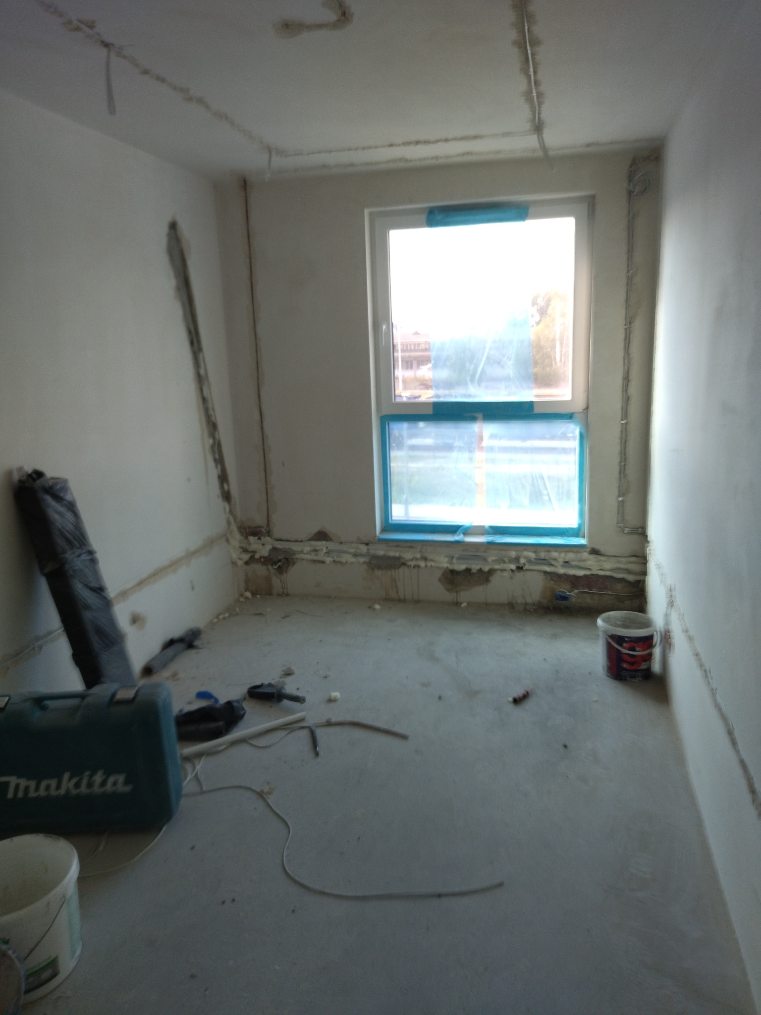 Wnętrze w trakcie prac remontowych: przewody elektryczne pod sufitem, zaklejone okno, narzędzia i wiaderka na podłodze. Widoczne ślady po kuciu w ścianach.