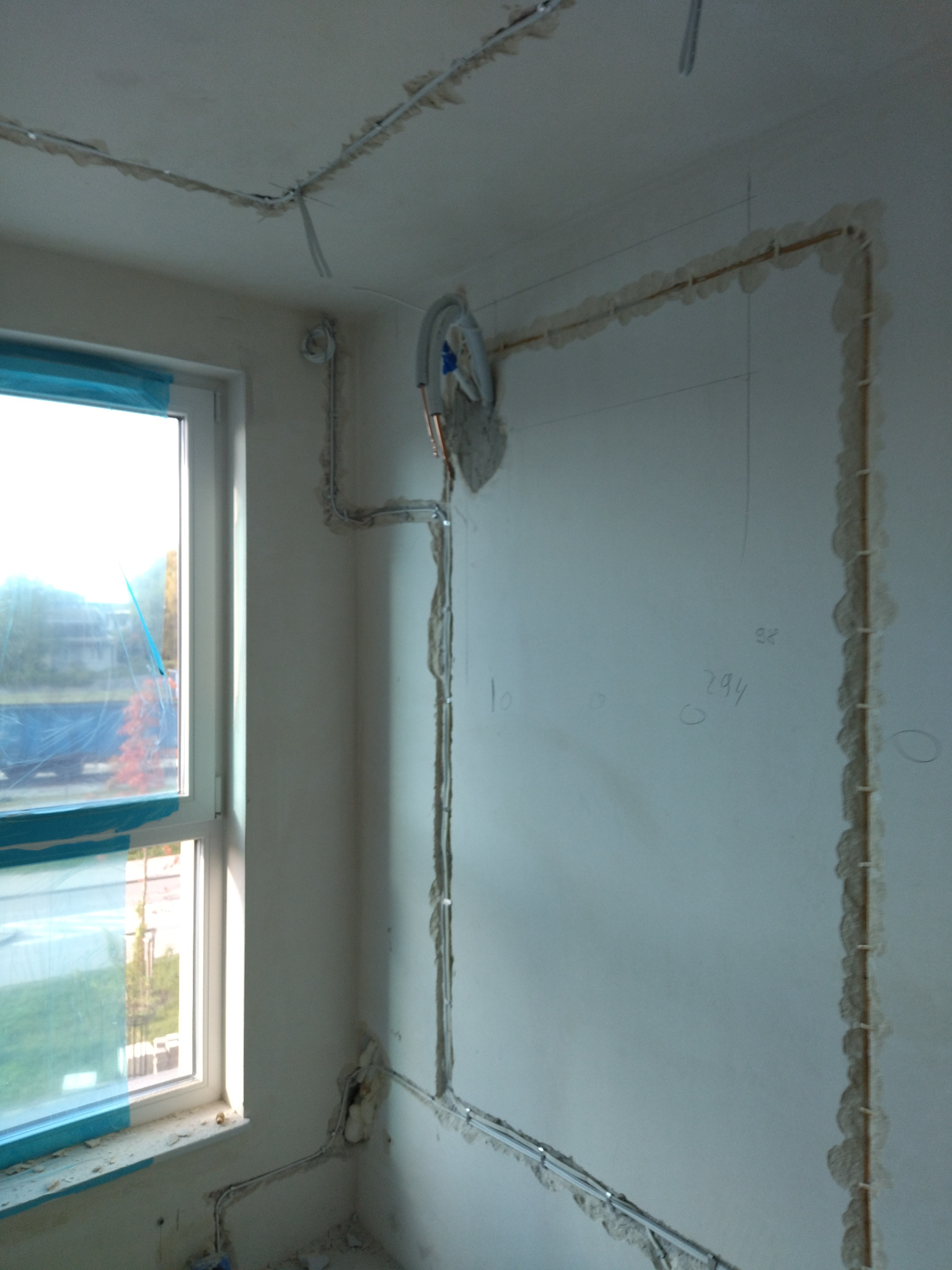 Białe kable elektryczne w bruzdach na ścianie i suficie pokoju w trakcie remontu, widoczne oznaczenia na ścianie, okno z niebieską folią ochronną.