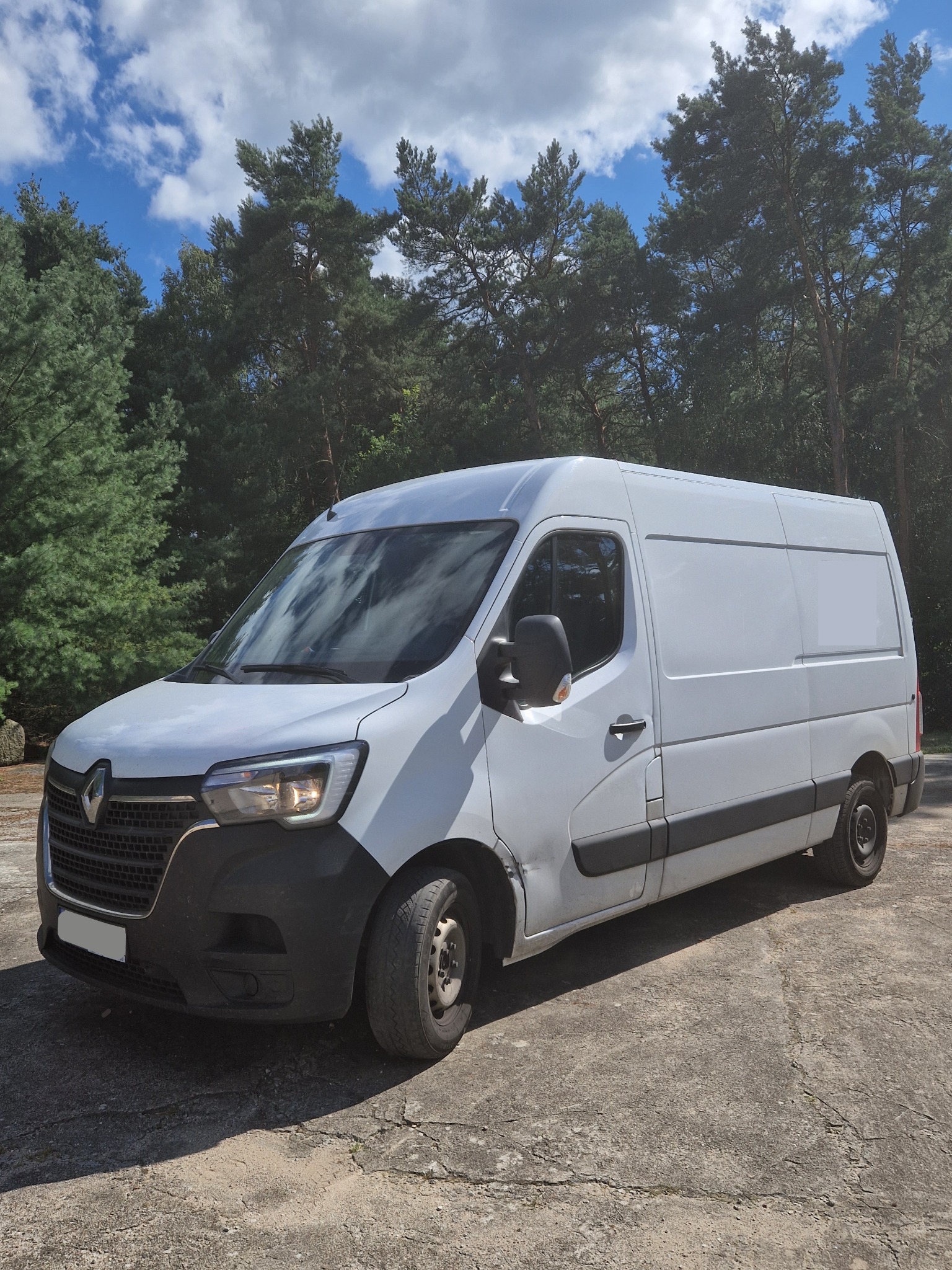 Biały Renault Master zaparkowany na betonowym placu, zarysowany błotnik, w tle las iglasty i błękitne niebo z chmurami.