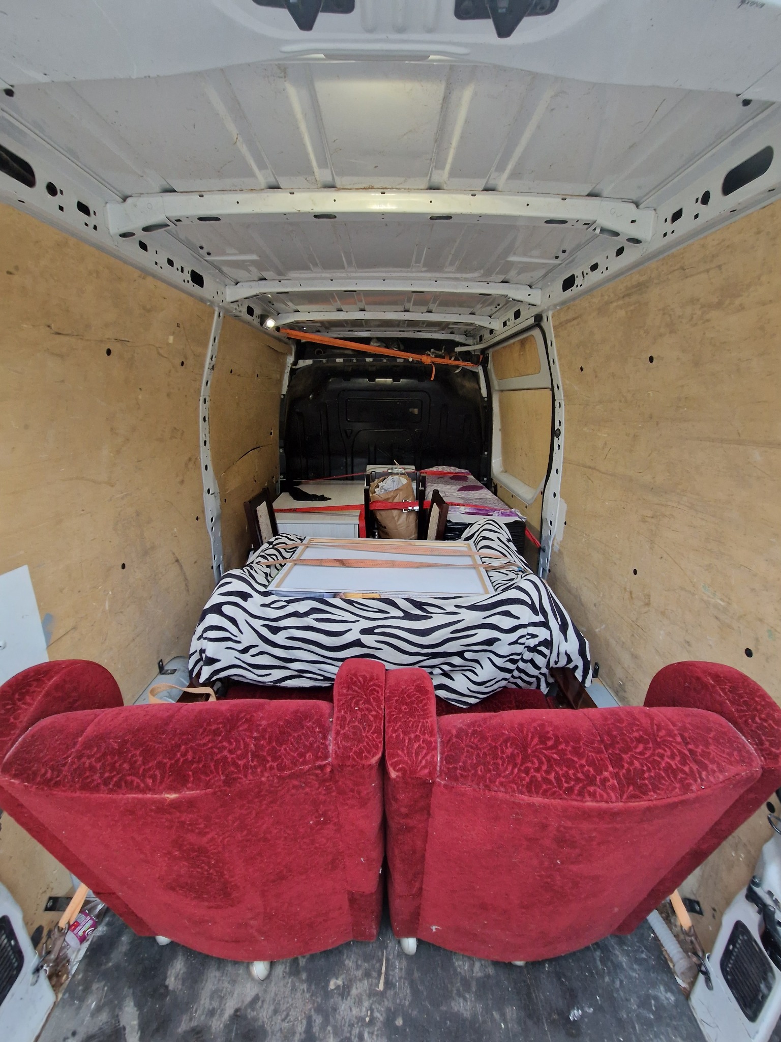 Wnętrze furgonetki wypełnione meblami, w tym czerwonym fotelem z ozdobnym wzorem i obrazem przykrytym kocem w zebrę, zabezpieczone pasami transportowymi.