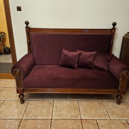 Ester-Tap - Burgundowa sofa z drewnianą ramą i dwoma poduszkami na tle beżowych płytek podłogowych i widocznym fragmentem pokoju z wiklinowym fotelem.