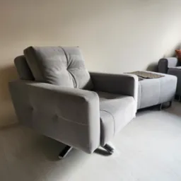 Szary, tapicerowany fotel obrotowy z podnóżkiem i dekoracyjną podkładką na podłodze z paneli w jasnym wnętrzu.