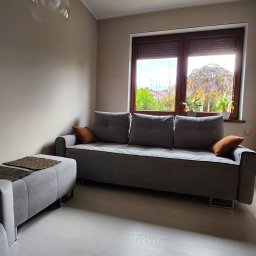 Ester-Tap - Szara sofa z poduszkami i pufą w salonie w Słubicach, widok na ogród przez okno z roletami.