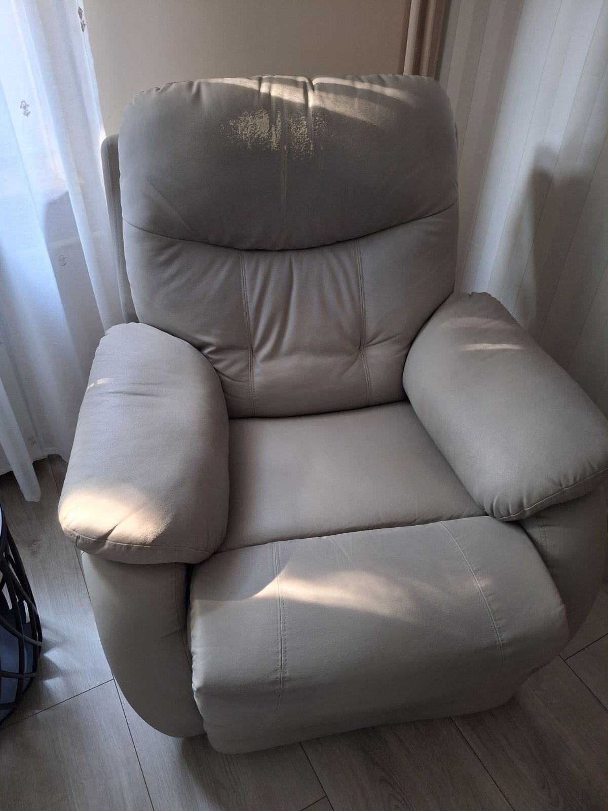 Jasnoszary fotel z funkcją relaks z widocznymi śladami użytkowania na zagłówku, stojący na drewnianej podłodze przy zasłoniętym oknie.
