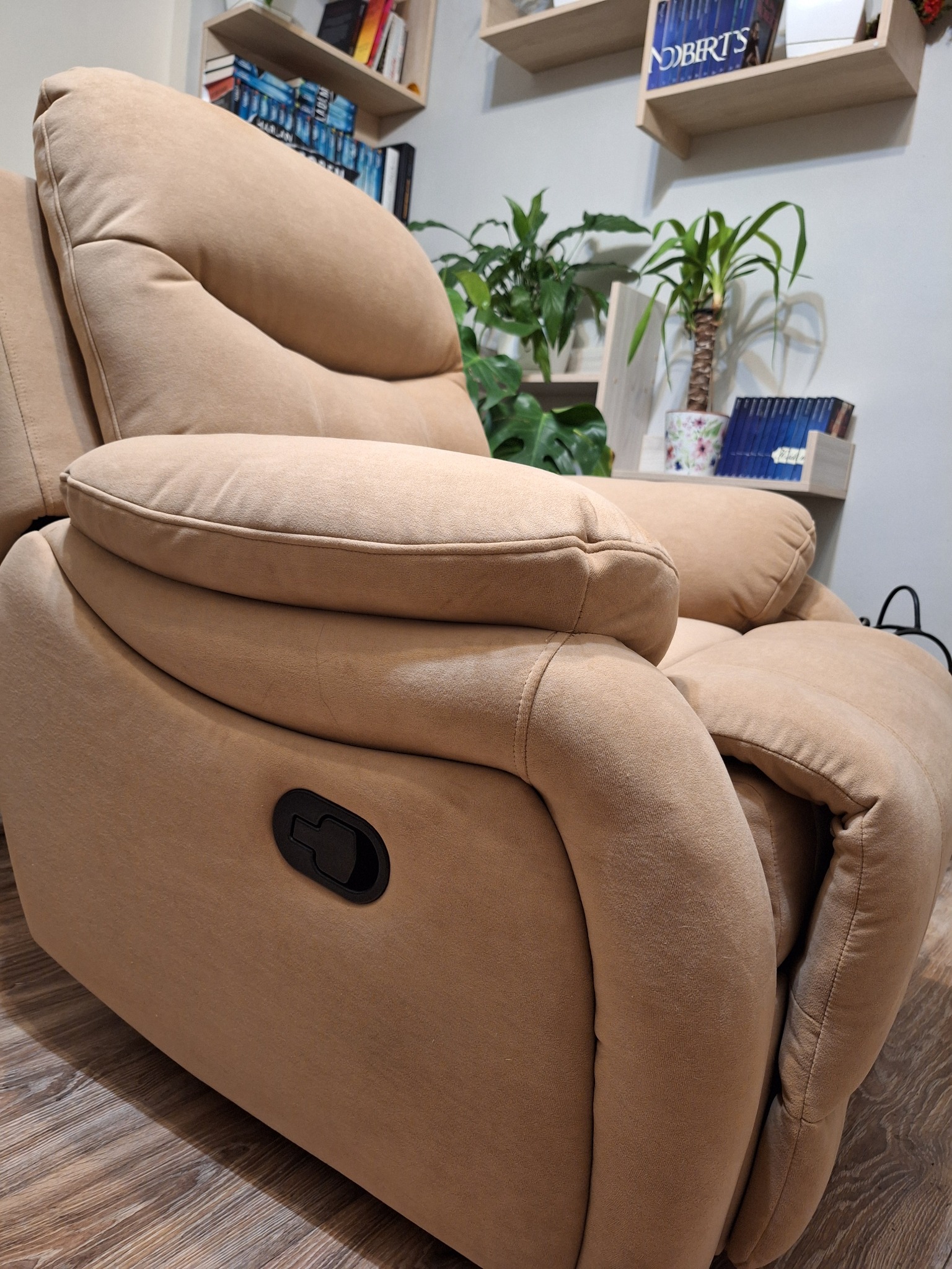 Beżowy fotel tapicerowany z funkcją relaksacyjną, widoczny mechanizm zmiany pozycji, w tle półki z książkami i roślinami doniczkowymi.