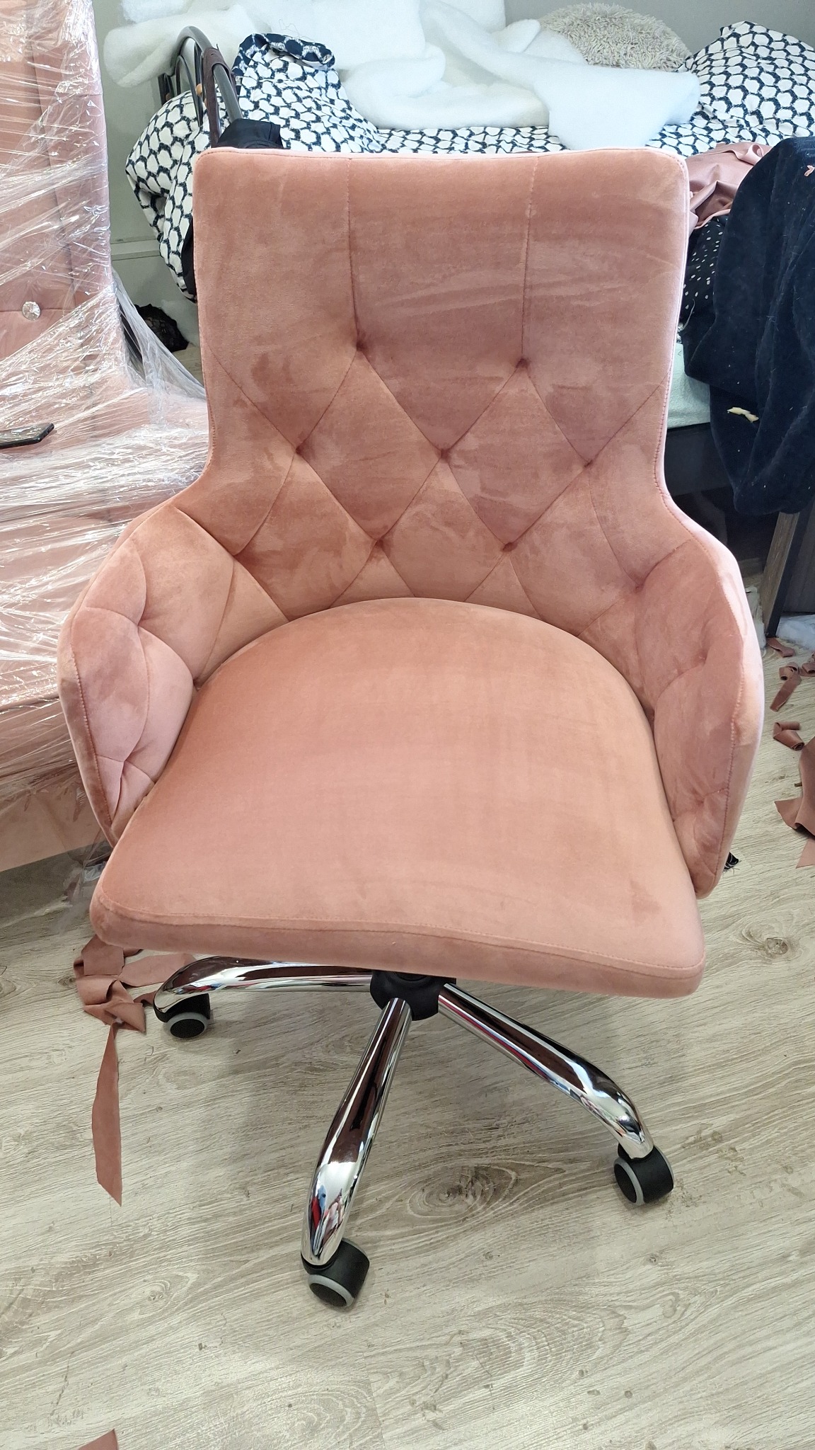 Różowe, tapicerowane krzesło biurowe na kółkach z pikowanym oparciem, wykonane w Słubicach, stoi na podłodze obok skrawków materiału i innego mebla owiniętego folią.