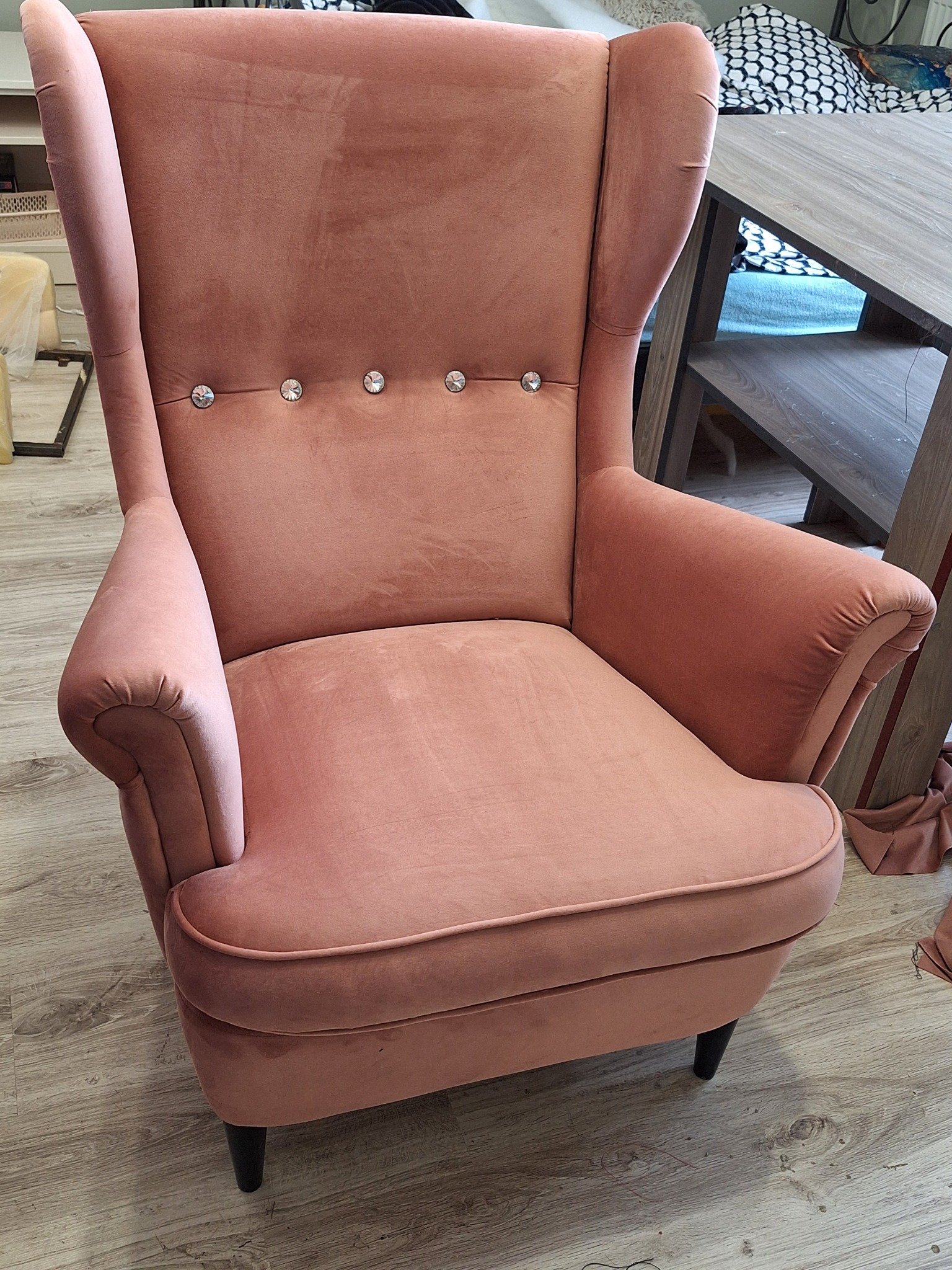 Różowy, welurowy fotel typu uszak z ozdobnymi kryształkami na oparciu, stojący na podłodze z paneli.