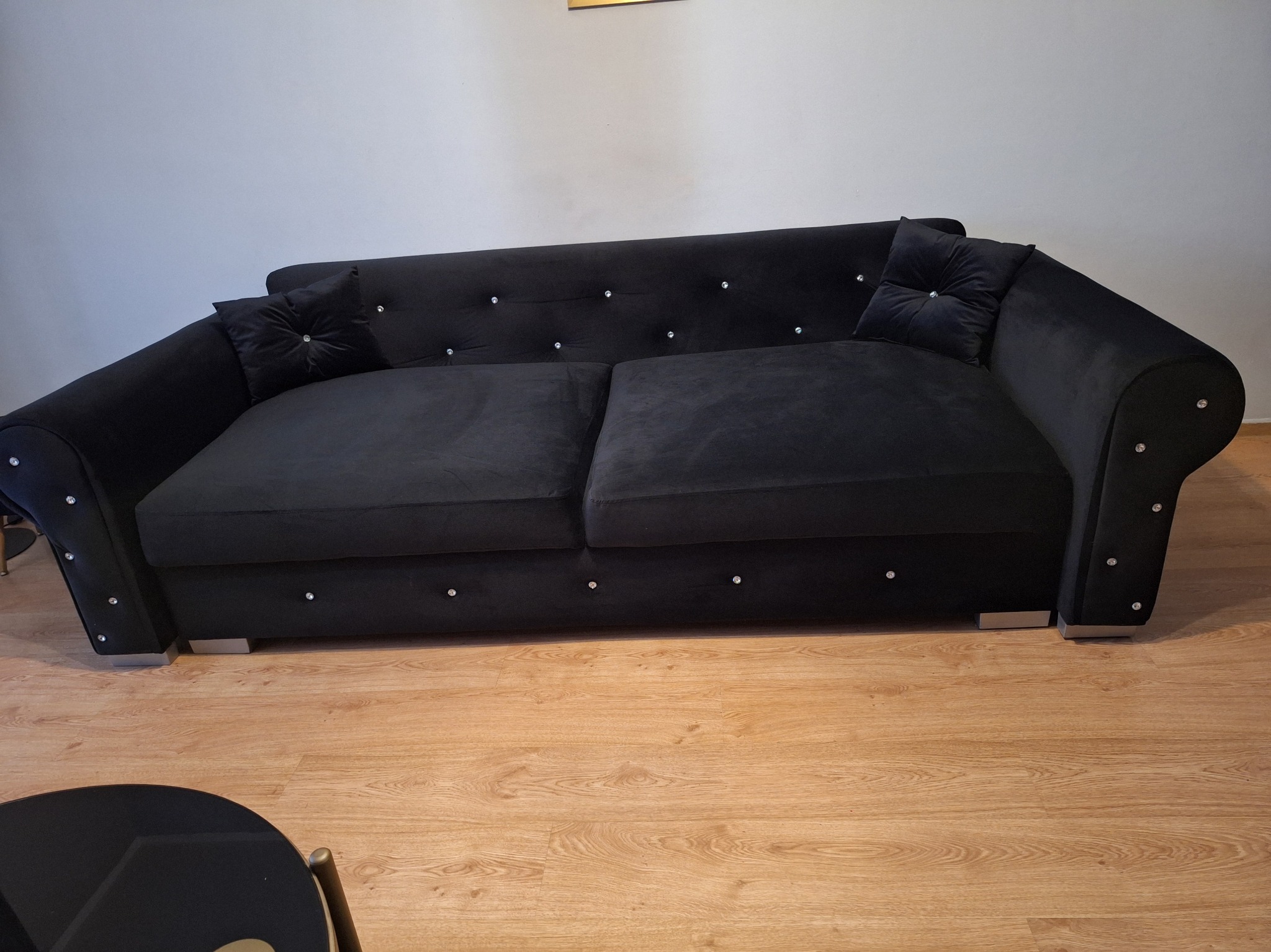 Czarna sofa z weluru z dekoracyjnymi kryształkami na oparciu i bokach, z dwoma poduszkami, stojąca na drewnianej podłodze. Widoczny fragment czarnego stolika kawowego.
