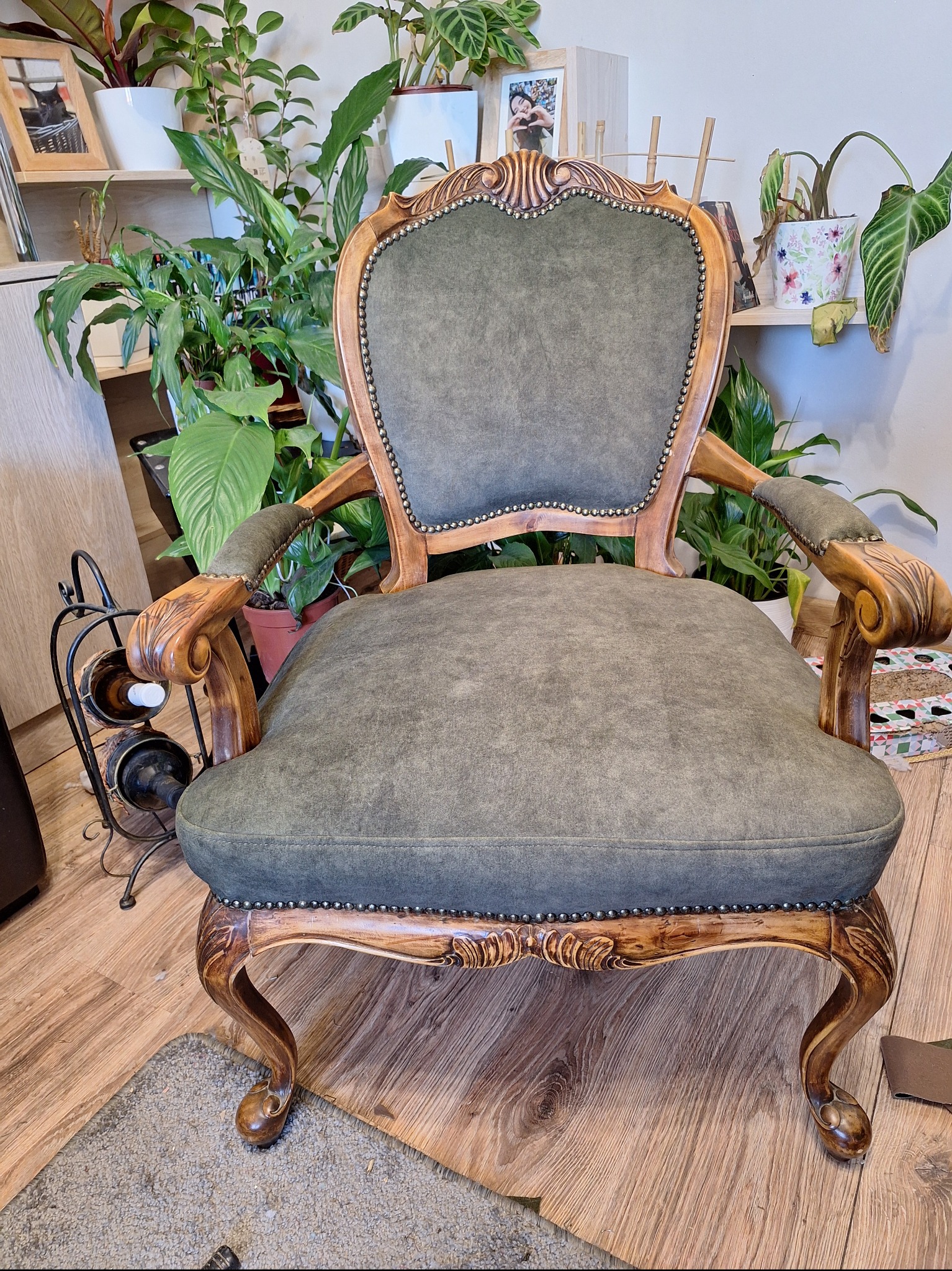 Odrestaurowany fotel w stylu vintage z zieloną tapicerką i rzeźbionymi drewnianymi nogami, otoczony roślinami doniczkowymi w jasnym wnętrzu.