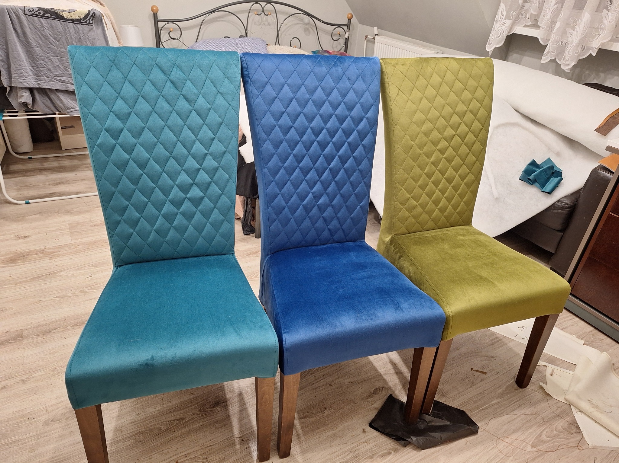 Trzy krzesła z wysokim oparciem, każde w innym kolorze: turkusowym, niebieskim i oliwkowym, z pikowaniem w romby, stojące obok siebie na podłodze.