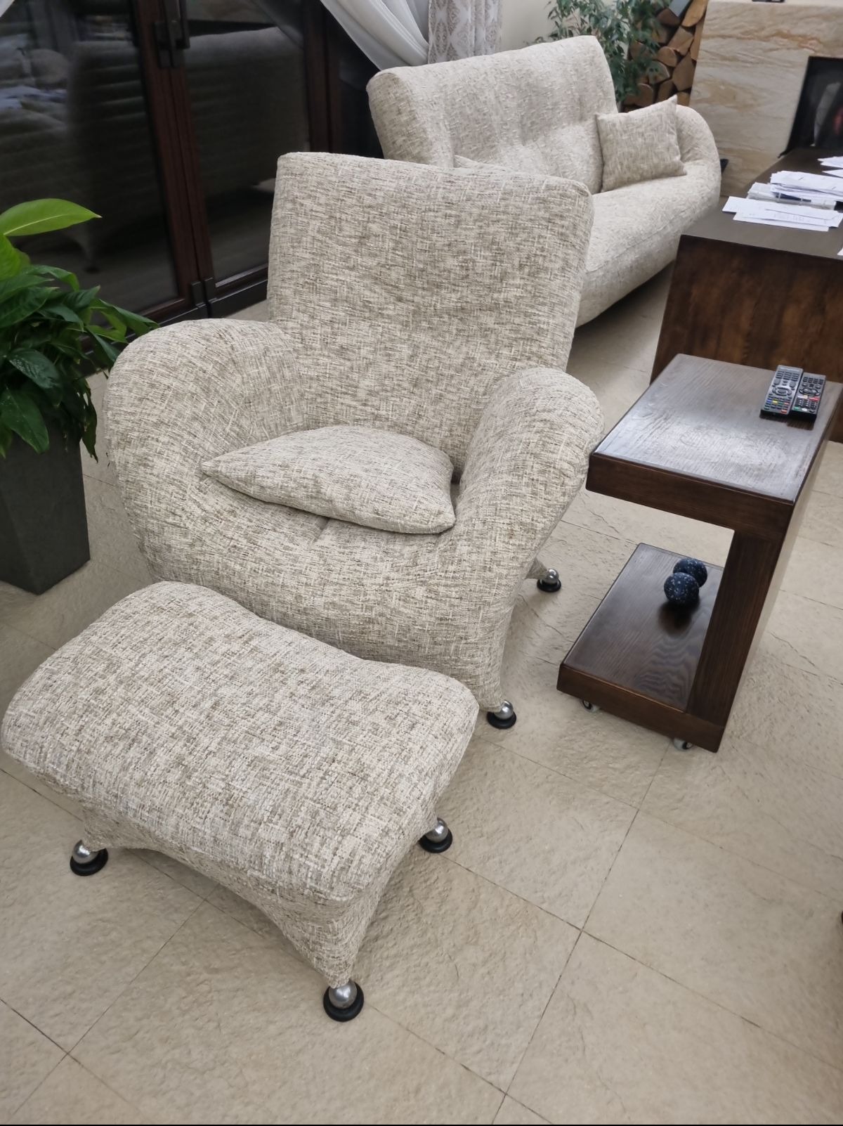 Tapicerowany fotel z podnóżkiem w jasnej, teksturowanej tkaninie, obok ciemny stolik z pilotami, w tle sofa w podobnej stylistyce.