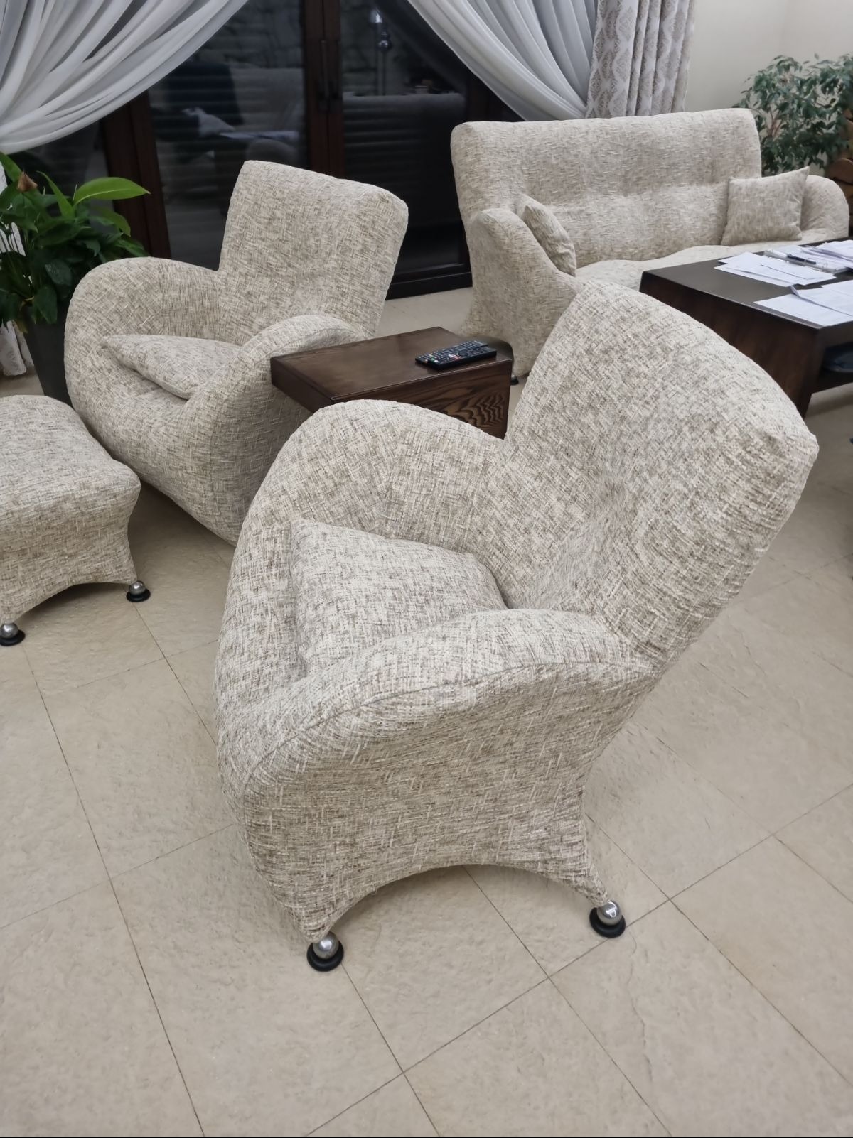 Komplet mebli tapicerowanych w jasnej, teksturowanej tkaninie: dwa fotele z poduszkami, sofa i pufa na nóżkach z kółkami, ustawione w salonie z ciemnymi stolikami kawowymi i zasłonami w tle.