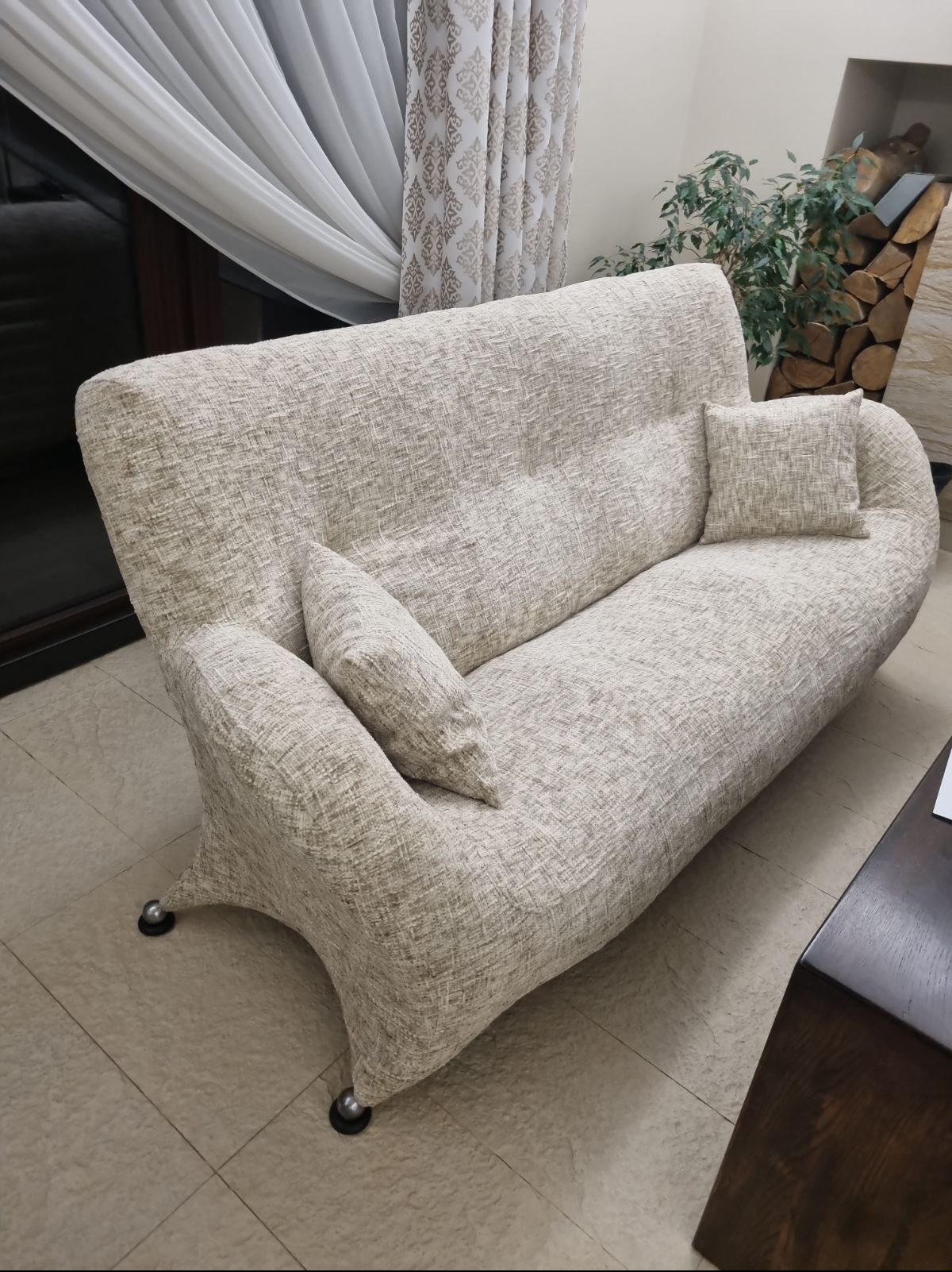 Beżowa sofa dwuosobowa z poduszkami, tapicerowana tkaniną o wyraźnej fakturze, na niskich nóżkach z metalowymi kulkami, w tle zasłony i dekoracyjny kominek z drewnem.