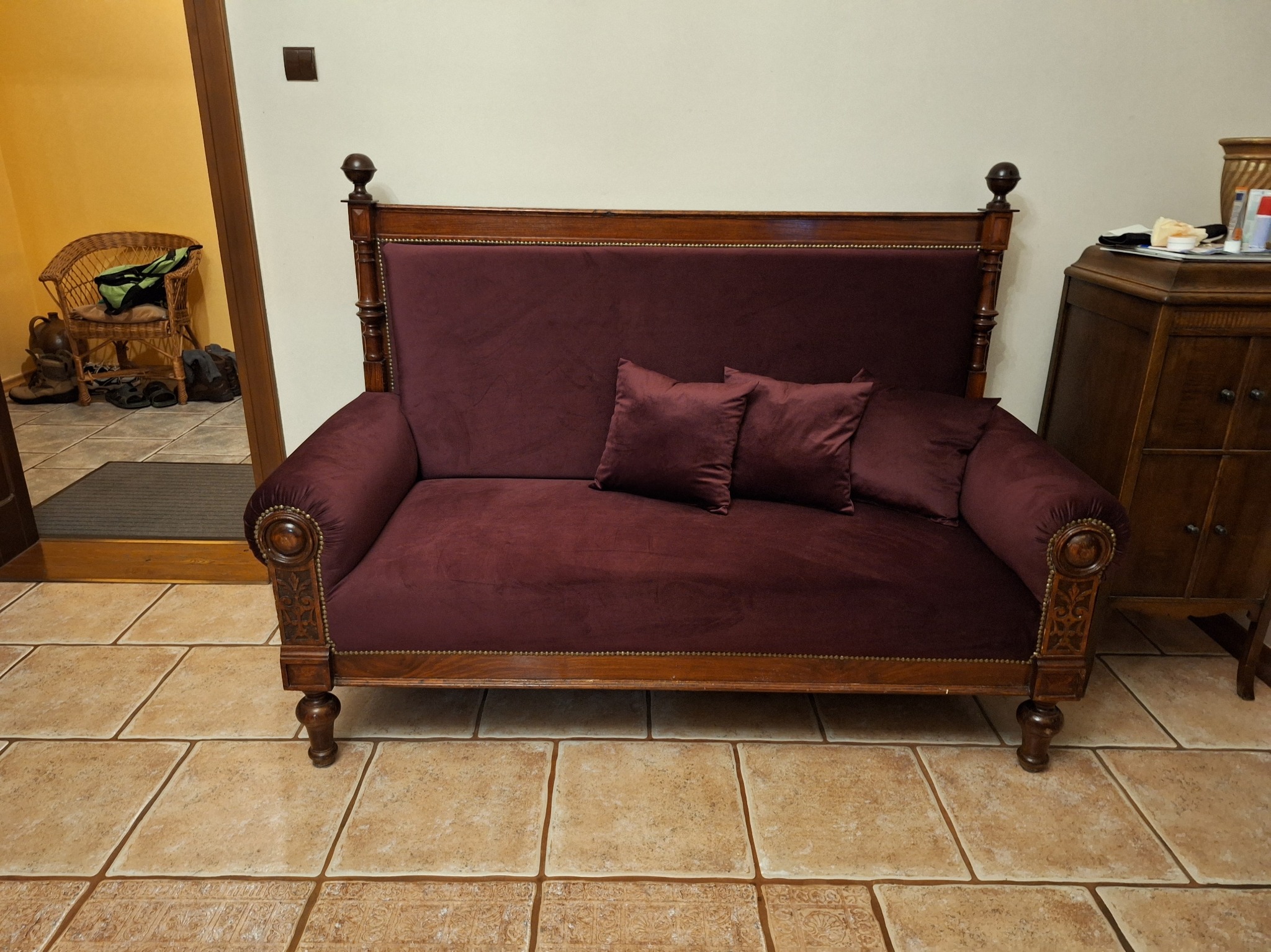 Burgundowa sofa z drewnianą ramą i dwoma poduszkami na tle beżowych płytek podłogowych i widocznym fragmentem pokoju z wiklinowym fotelem.