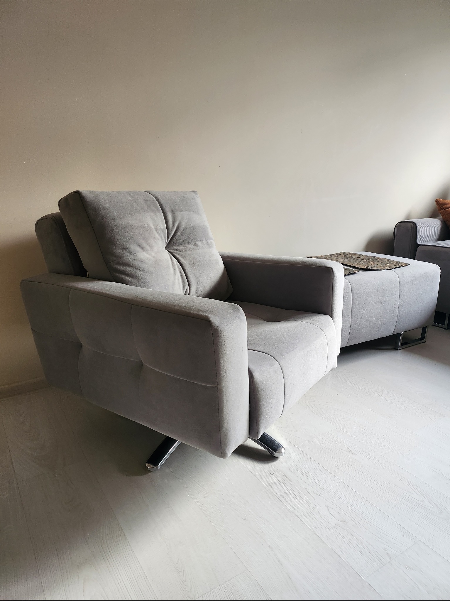 Szary, tapicerowany fotel obrotowy z podnóżkiem i dekoracyjną podkładką na podłodze z paneli w jasnym wnętrzu.