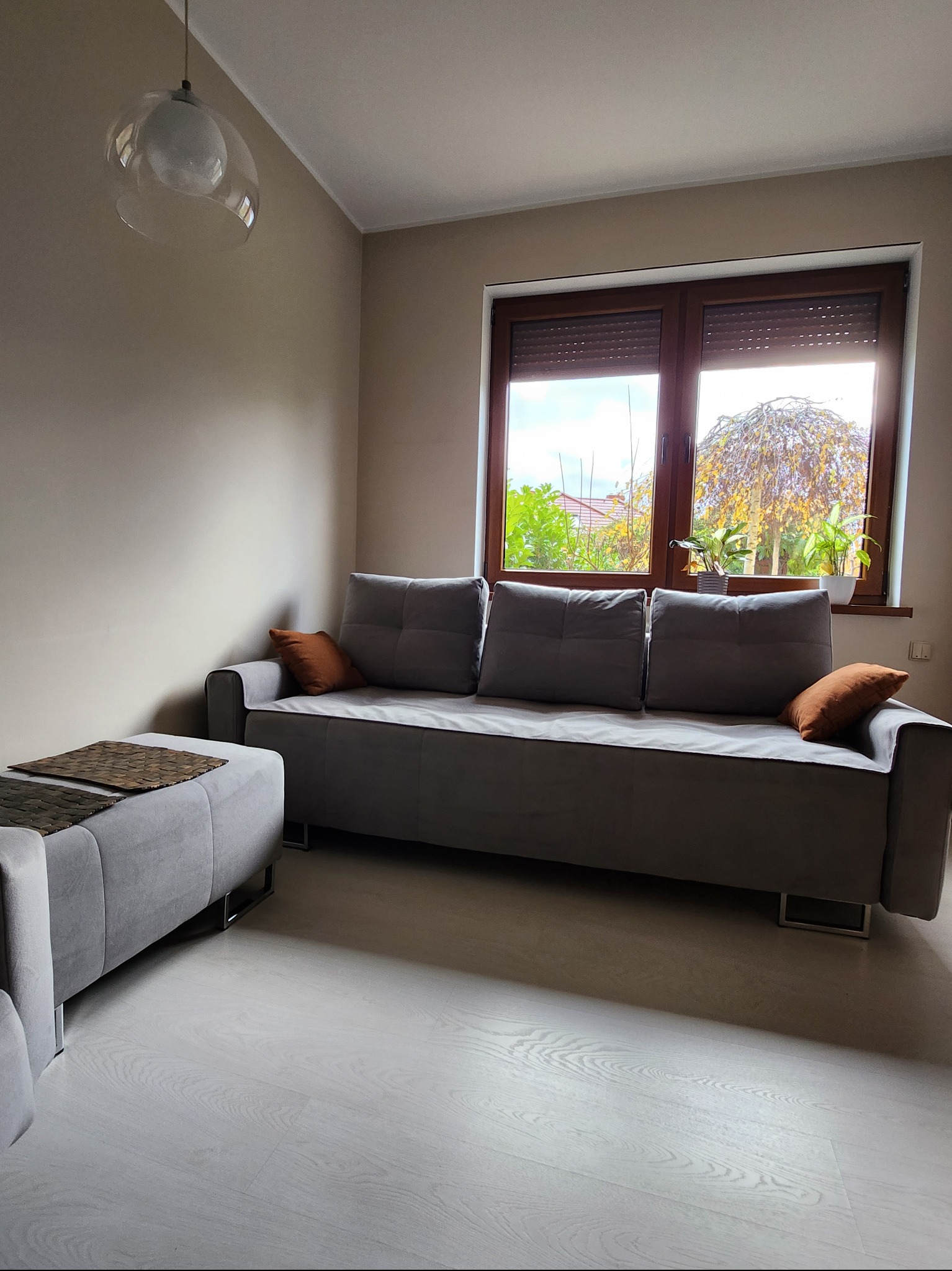 Szara sofa z poduszkami i pufą w salonie w Słubicach, widok na ogród przez okno z roletami.