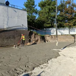 Pracownik w kamizelce odblaskowej wyrównuje świeżo wylaną, szarą posadzkę betonową przy użyciu wibratora powierzchniowego. W tle ściana budynku i drzewa.