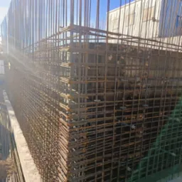 Siatka zbrojeniowa z pionowymi prętami wystającymi ponad poziom gruntu, widoczne elementy betonowe i budynek w tle.