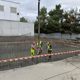 ŻELBETPOL SPÓŁKA Z OGRANICZONĄ ODPOWIEDZIALNOŚCIĄ - Wylewanie betonu do szalunku zbrojonego na budowie fundamentów, trzech pracowników w odblaskowych kamizelkach obsługuje pompę do betonu, widoczne zbrojenie i szalunek zabezpieczony taśmą ostrzegawczą.