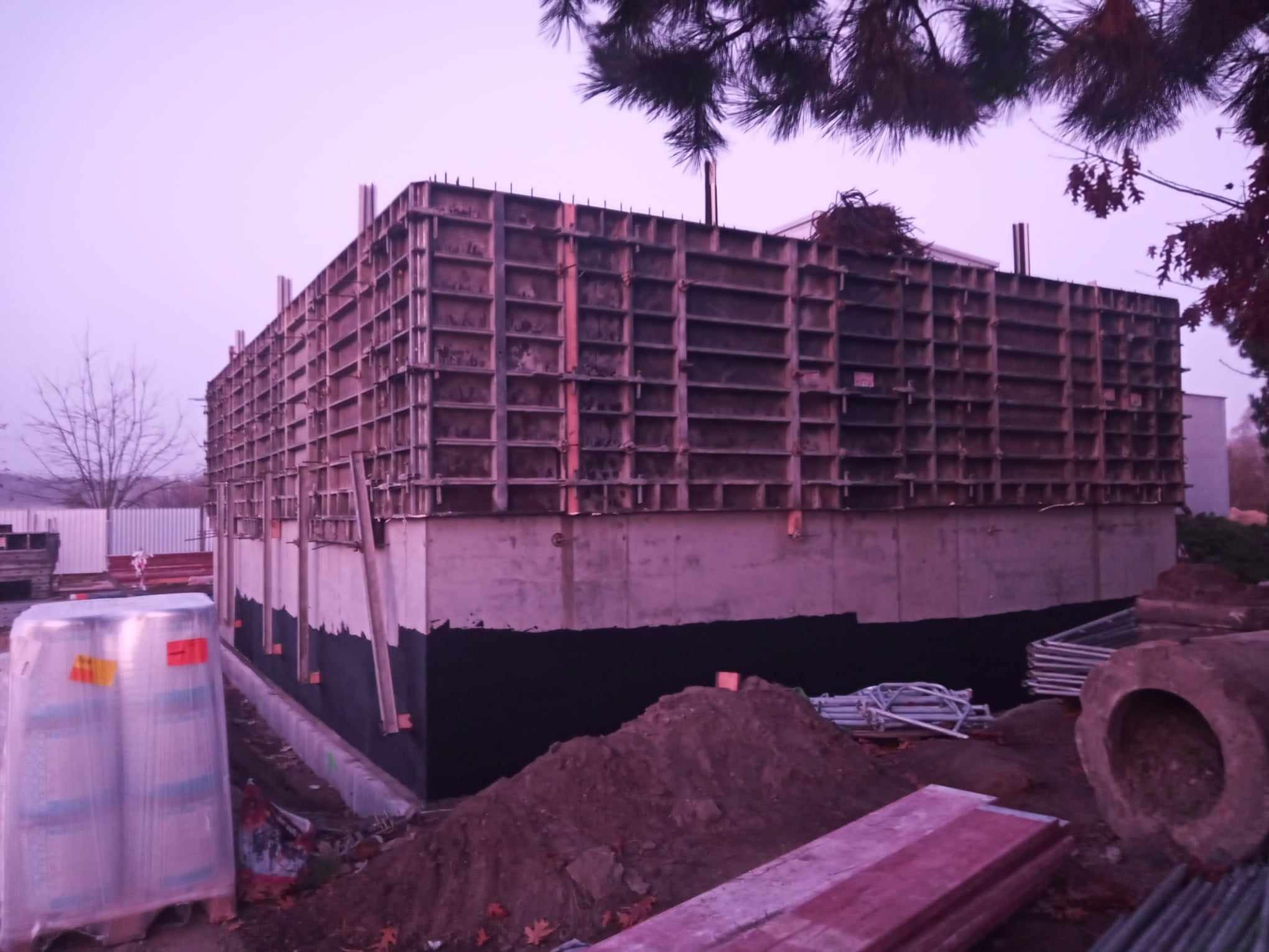 Szare betonowe ściany budynku w trakcie budowy z widocznym szalunkiem i izolacją fundamentów, w tle drzewa i pochmurne niebo.