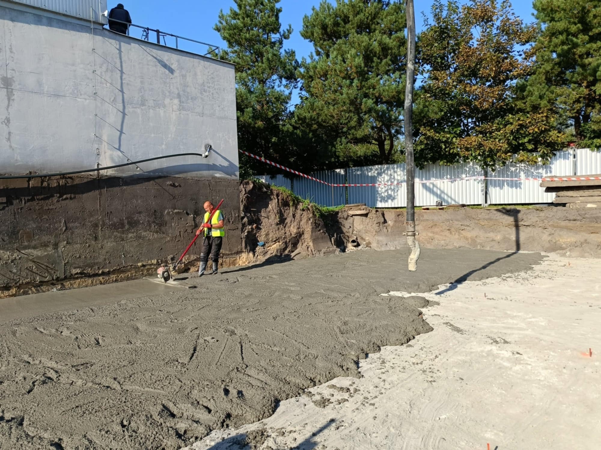 Pracownik w kamizelce odblaskowej wyrównuje świeżo wylaną, szarą posadzkę betonową przy użyciu wibratora powierzchniowego. W tle ściana budynku i drzewa.