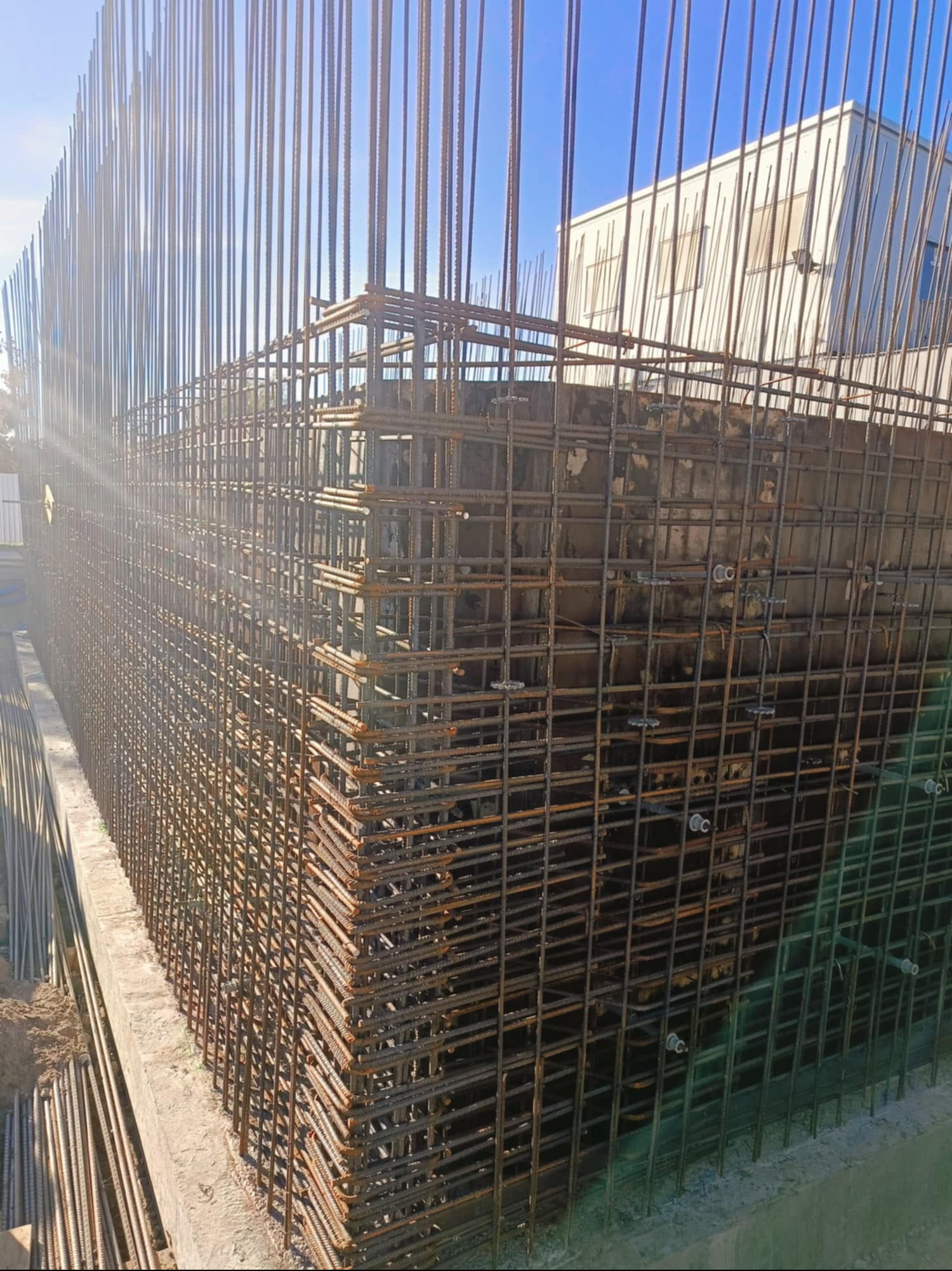 Siatka zbrojeniowa z pionowymi prętami wystającymi ponad poziom gruntu, widoczne elementy betonowe i budynek w tle.