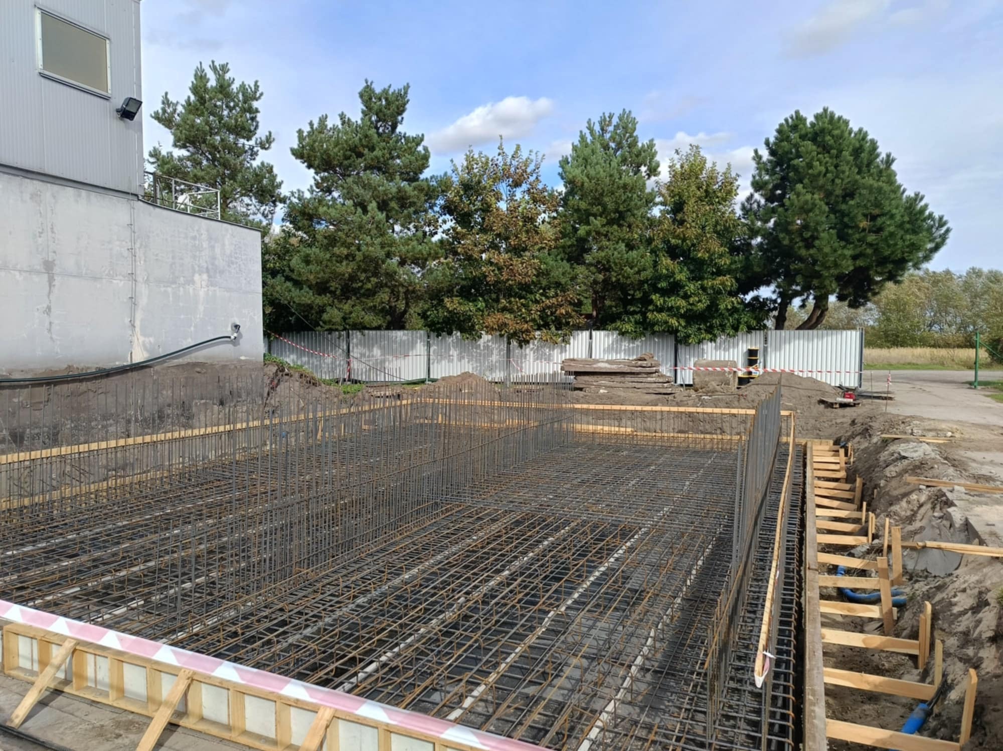 Siatka zbrojeniowa w wykopie fundamentowym, przygotowana do zalania betonem, z widocznymi szalunkami i elementami instalacji, w tle budynek przemysłowy i drzewa.