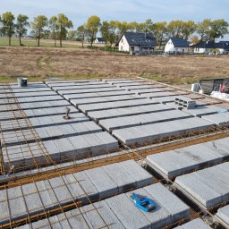 DAW- BUD - Wieniec stropowy z szalunkiem i zbrojeniem na budowie domu jednorodzinnego, widok z góry na pustaki szalunkowe i stalowe pręty, w tle zabudowa jednorodzinna i drzewa.