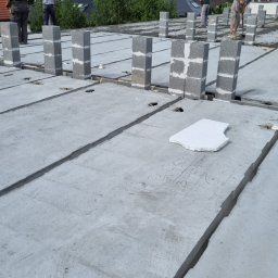 DAW- BUD - Wznoszenie ścian nośnych z bloczków betonowych na betonowym stropie prefabrykowanym, widoczne zbrojenia, na dalszym planie pracownicy budowlani i zabudowa jednorodzinna.