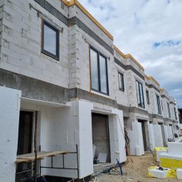 DAW- BUD - Szeregówka w stanie surowym otwartym z częściowo zamontowaną stolarką okienną i przygotowanymi otworami garażowymi obłożonymi styropianem, widoczne materiały budowlane na terenie budowy.