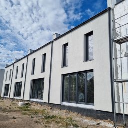 DAW- BUD - Rząd szeregowych domów jednorodzinnych w trakcie wykańczania elewacji, jeden z domów z widocznym rusztowaniem i nieotynkowaną ścianą, pozostałe z białą elewacją i ciemnymi oknami, błękitne niebo...