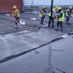 DAW- BUD - Ekipa budowlana w żółtych kamizelkach i kaskach, w trakcie wylewania i rozprowadzania betonu na zbrojonej powierzchni dachu budynku w szary, pochmurny dzień.