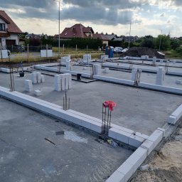 DAW- BUD - Zalany fundament z bloczków betonowych, wystające pręty zbrojeniowe, widoczne zarysy przyszłych ścian, w tle zabudowa jednorodzinna i pochmurne niebo.