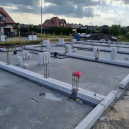Zalany fundament z bloczków betonowych, wystające pręty zbrojeniowe, widoczne zarysy przyszłych ścian, w tle zabudowa jednorodzinna i pochmurne niebo.