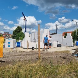 DAW- BUD - Budowa domu z białych bloczków silikatowych, widoczny dźwig, dwóch pracowników przy ścianie, palety z materiałem budowlanym i baner reklamowy w tle błękitne niebo z chmurami.