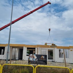 DAW- BUD - Budowa domu z białych bloczków: czerwony dźwig podnosi element konstrukcyjny, widoczny baner reklamowy 'ANNA Nieruchomości' oraz drabina oparta o ścianę.