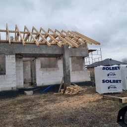 DAW- BUD - Surowy stan domu w trakcie budowy: ściany z białych bloczków, drewniana konstrukcja dachu, paczki materiałów budowlanych Solbet z logo producenta na działce.